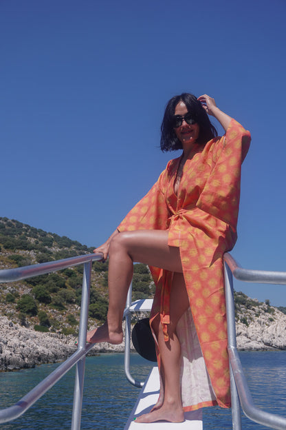 A woman wearing a bright orange and yellow patterned kaftan-style cover-up sits on the bow of a boat, with blue water and rocky shoreline in the background.