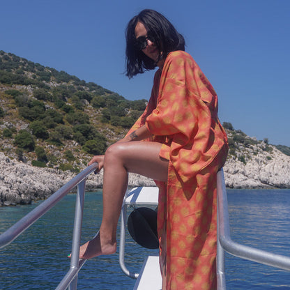 A woman wearing a loose orange patterned kaftan-style dress sits on the white deck railing of a boat, with blue water and a green rocky shoreline in the background.