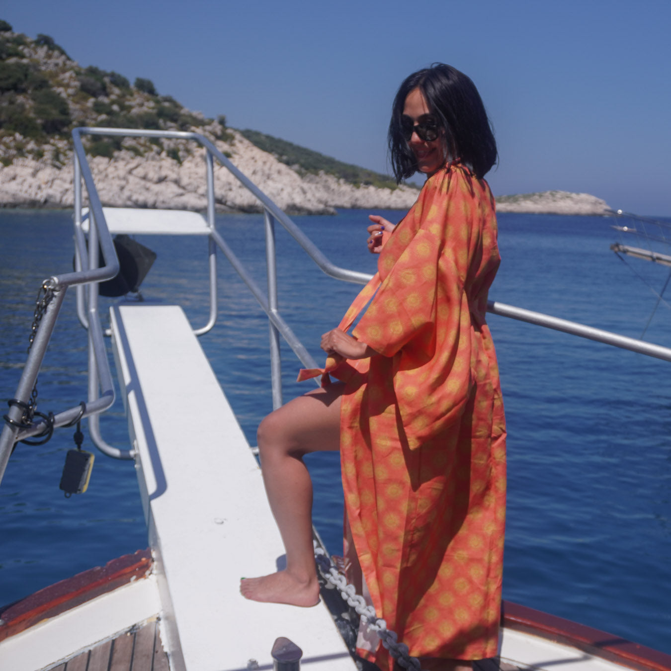 A woman wearing a bright orange sun patterned kaftan-style cover-up and sunglasses stands on the ramp of a boat with blue sea and rocky shoreline in the background.