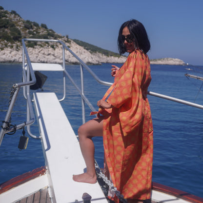 A woman wearing a bright orange sun patterned kaftan-style cover-up and sunglasses stands on the ramp of a boat with blue sea and rocky shoreline in the background.