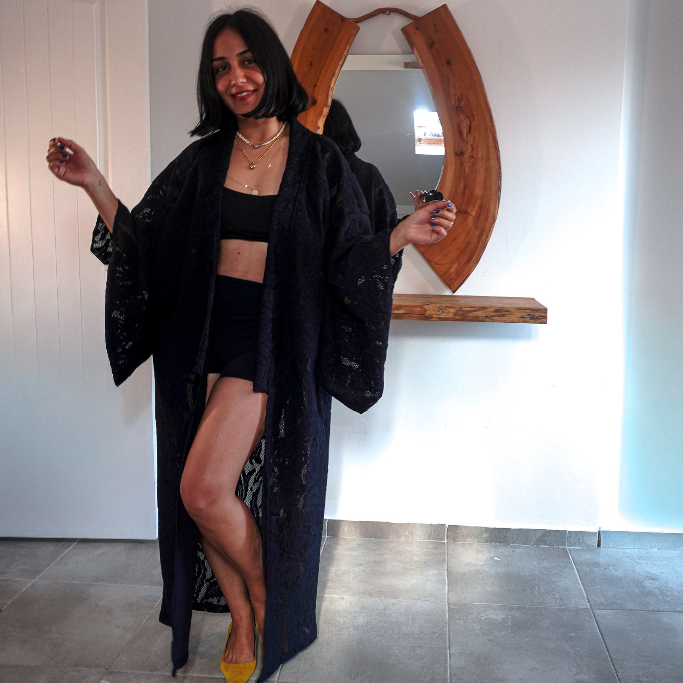 A woman stands indoors wearing a long black open-front robe with wide sleeves over a black crop top and black shorts, paired with yellow heels. A large wooden-framed mirror and white wall are behind her.