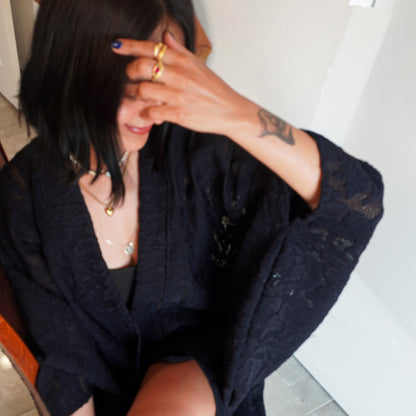 A woman wearing a black lace-trimmedr kimono over a black top, seated indoors with her hand covering part of her face. She is wearing multiple gold rings and layered gold necklaces.
