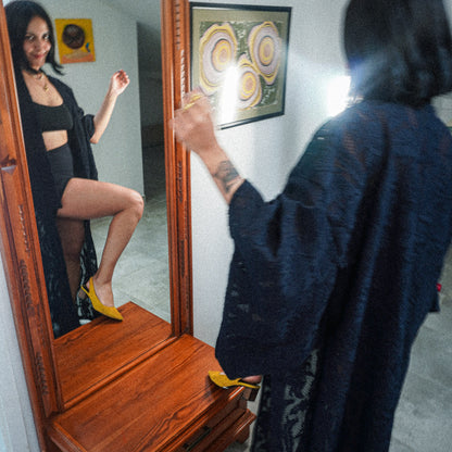 A woman stands in front of a wooden mirror, wearing a black lace kimono and yellow high heels, while her reflection shows her posing in a black outfit and the same yellow heels. The setting appears to be an indoor room with framed wall art and a gray floor.