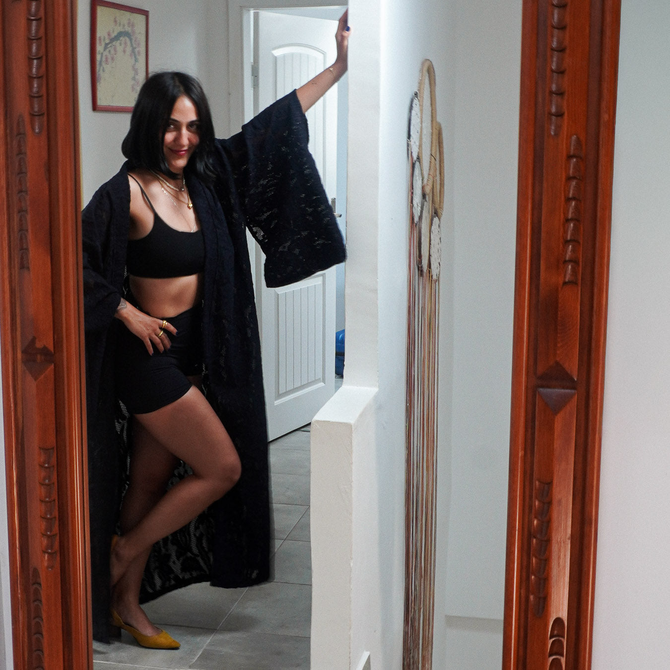 A woman posing in a doorway wearing a black lingerie set with a black open-front robe, framed by carved wooden door borders on both sides.
