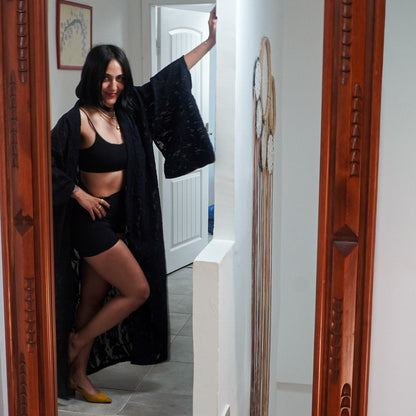 A woman posing in a doorway wearing a black lingerie set with a black open-front robe, framed by carved wooden door borders on both sides.