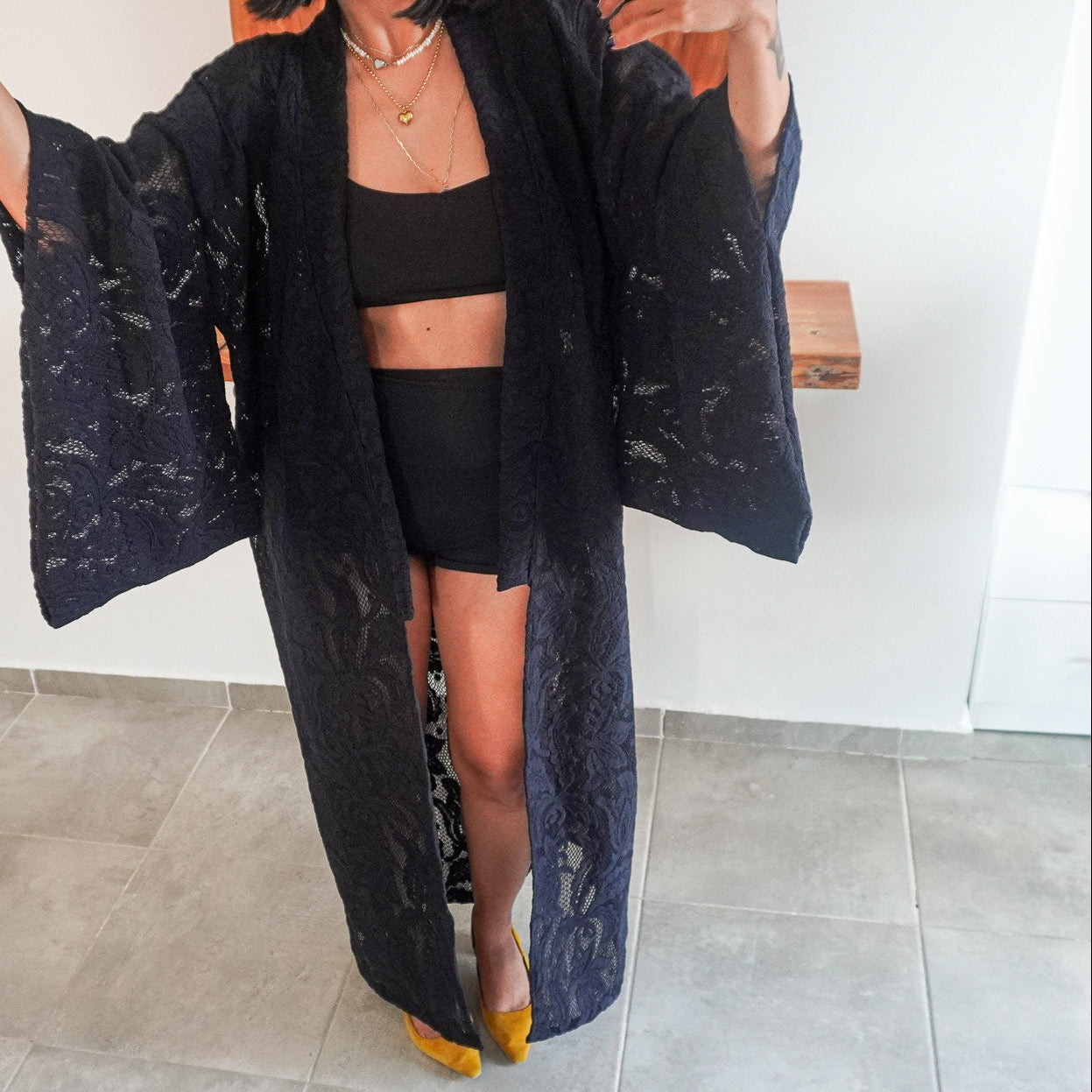 A woman modeling a long black lace kimono-style cardigan over a black crop top and shorts, standing indoors on a gray tiled floor against a white wall.