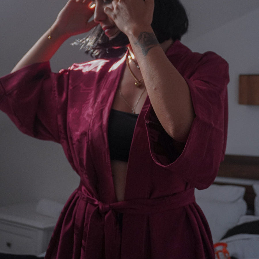 A woman wearing a deep burgundy satin  kimono over a black bralette, posing in a dimly lit bedroom.